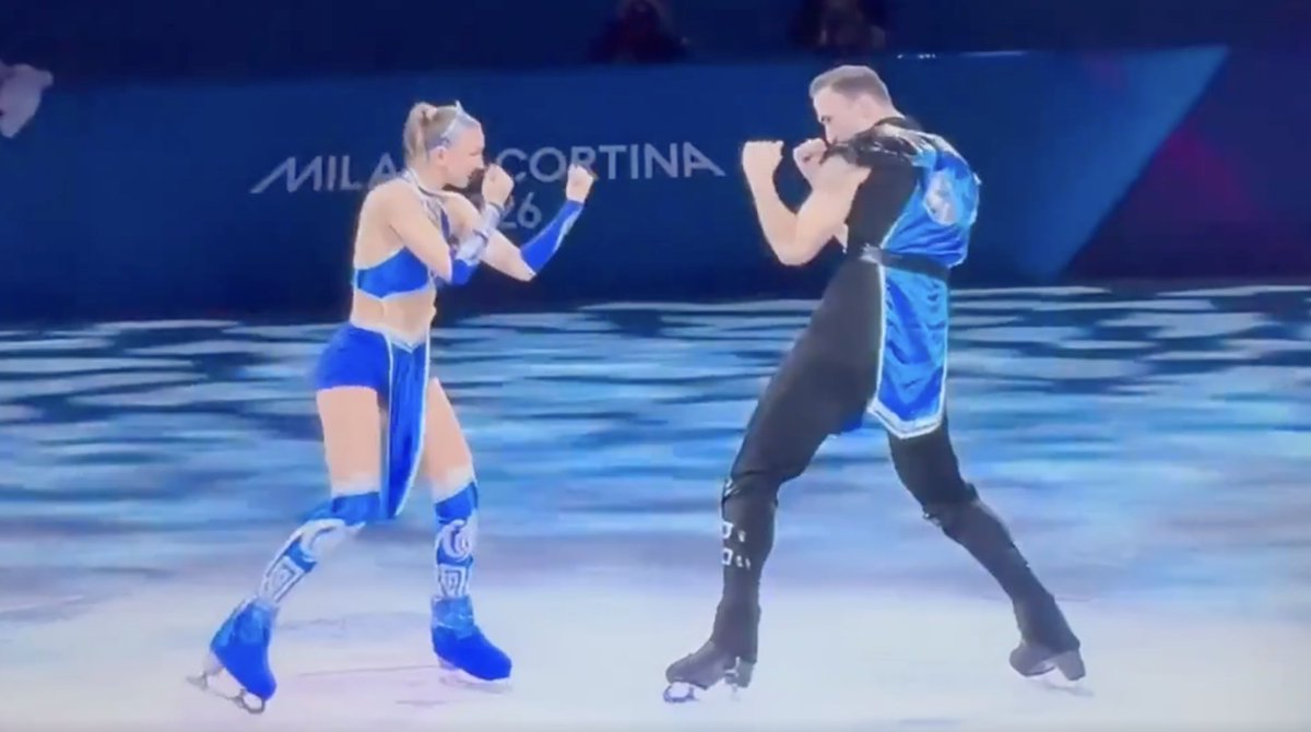 Two figure skaters dressed in blue Mortal Kombat-inspired costumes performing a combat-style routine on Olympic ice.