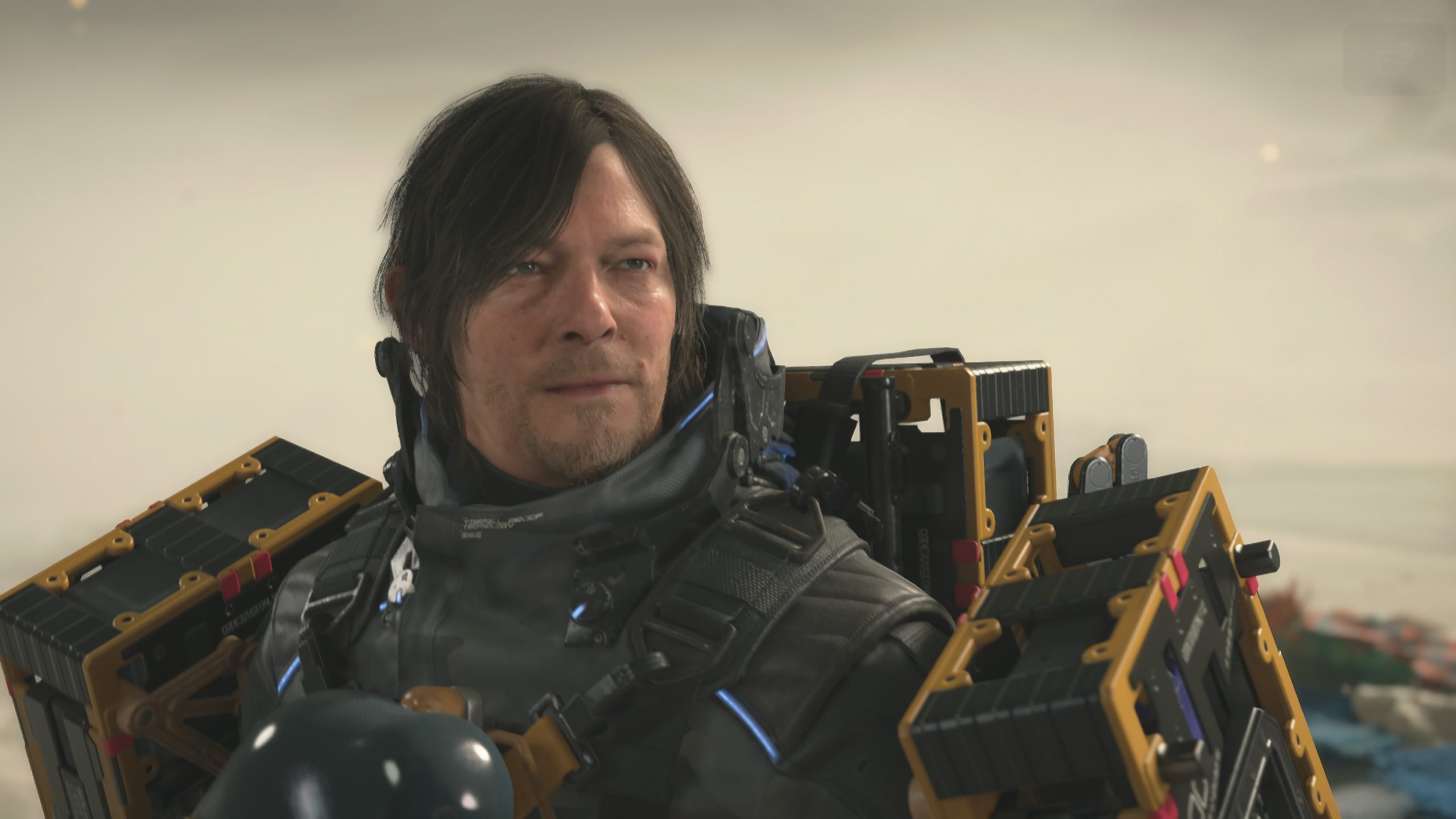 Gameplay image from Death Stranding 2: On the Beach showing Sam Porter Bridges in futuristic delivery gear carrying cargo equipment in a cinematic desert environment.